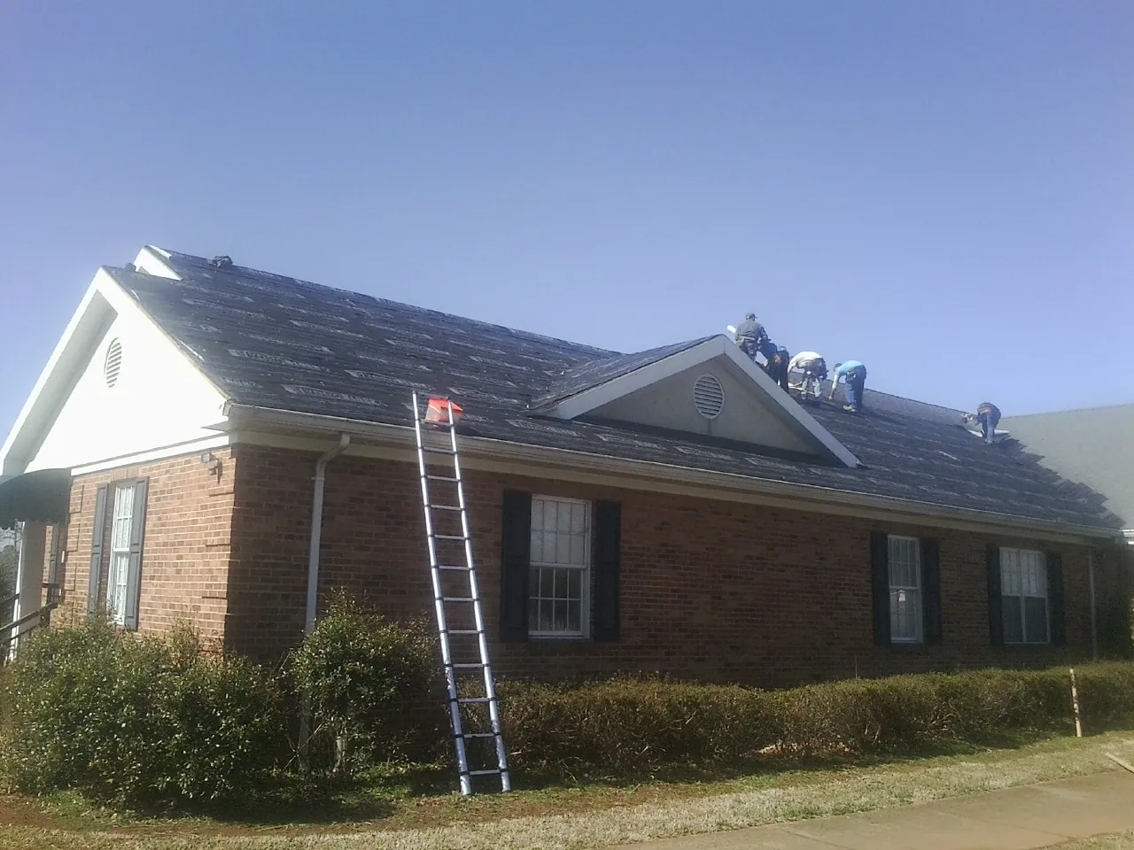 Crew tearing shingles off a brick ranch roof with a ladder set — Emergency Roof Repair work by our crew in Clifton Park