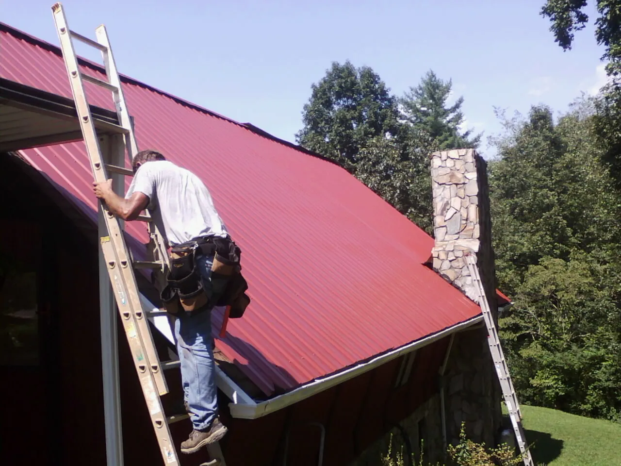 Roofer on a ladder beside a red metal roof and stone chimney — Soffit & Fascia Repair by Roofing Brothers in Clifton Park